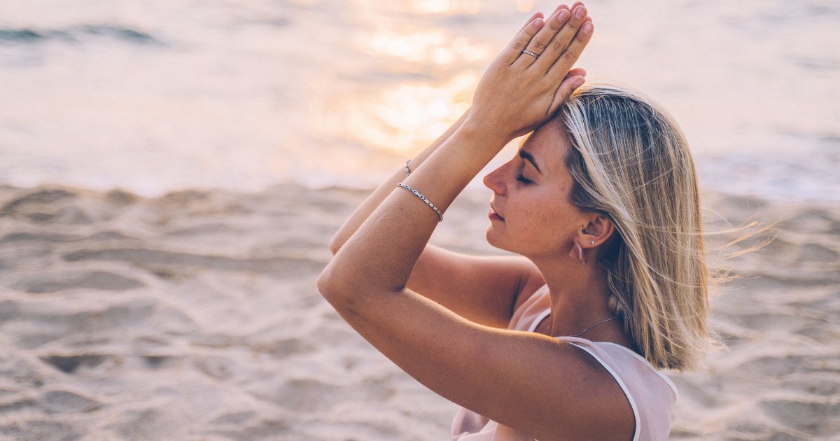 Eine Frau sitzt meditierend im Schneidersitz am Strand, mit Blick aufs Meer – umgeben von Ruhe, Natur und sanftem Licht.